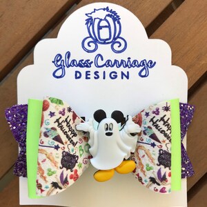 May include: A white and green bow with a purple glitter accent. The bow features a white ghost Mickey Mouse character with yellow shoes. The bow has a Halloween themed fabric with the words "Happy Halloween!" and "Hocus Pocus".