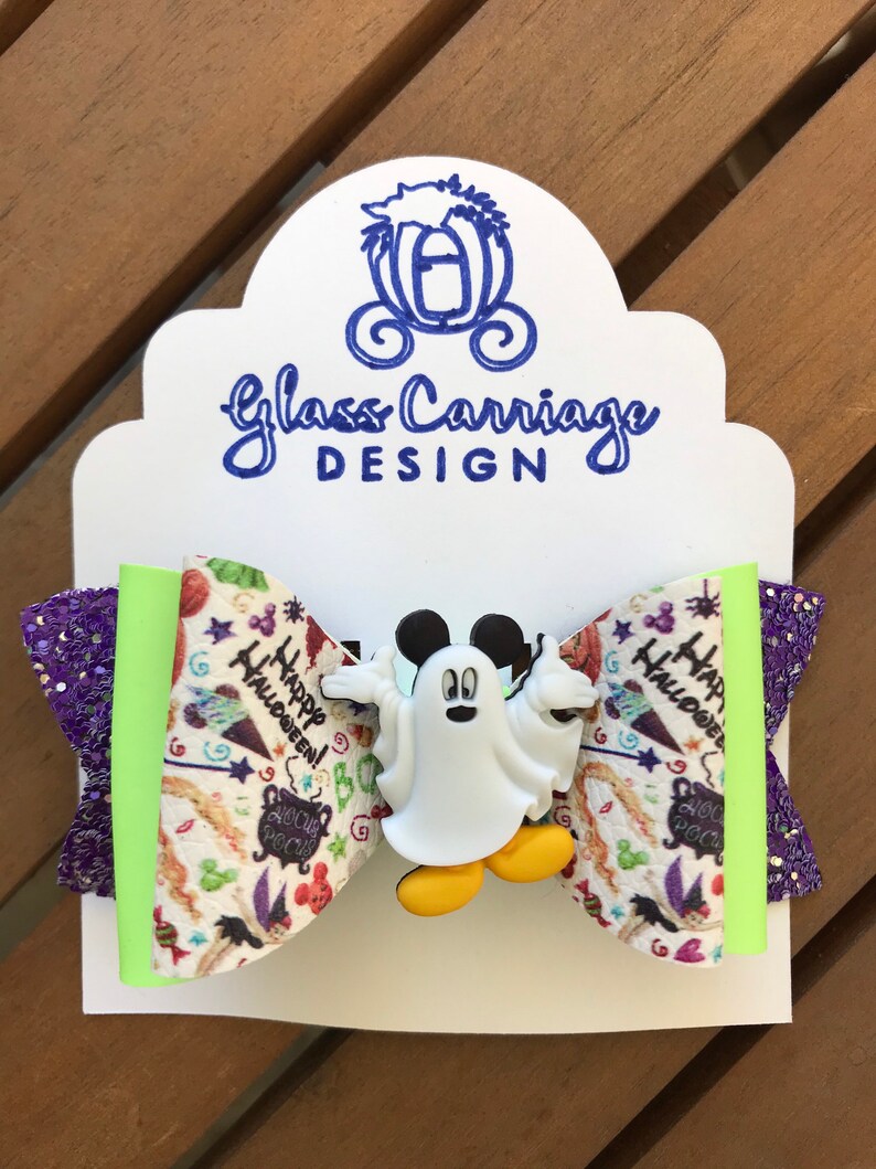 May include: A white and green bow with a purple glitter accent. The bow features a white ghost Mickey Mouse character with yellow shoes. The bow has a Halloween themed fabric with the words "Happy Halloween!" and "Hocus Pocus".