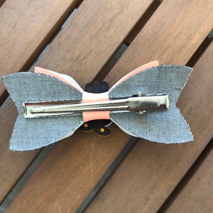 May include: A gray fabric bow with a peach colored center and a black and yellow butterfly design. The bow is attached to a silver alligator clip.