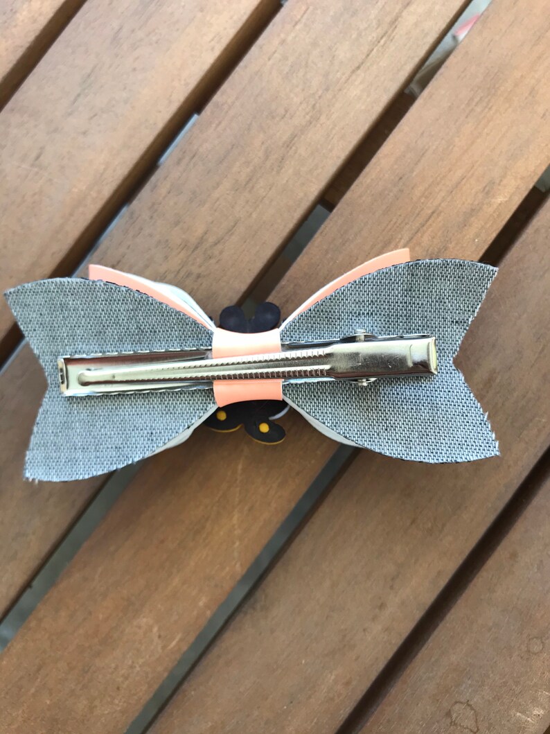 May include: A gray fabric bow with a peach colored center and a black and yellow butterfly design. The bow is attached to a silver alligator clip.