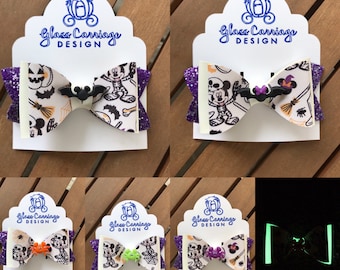Puede incluir: Un conjunto de cinco lazos para el pelo con temática de Halloween con un diseño de Mickey Mouse y Minnie Mouse. Los lazos están hechos de tela blanca con un estampado de Mickey Mouse y Minnie Mouse en blanco y negro. Los lazos tienen un ribete de purpurina morado y un amuleto de murciélago negro. Los lazos se muestran en tarjetas blancas con el texto "Glass Carriage Design". El último lazo se muestra brillando en la oscuridad.