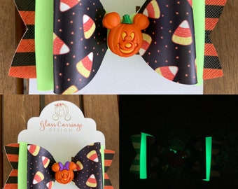 Puede incluir: Un lazo negro y verde con un diseño que brilla en la oscuridad con maíz dulce y una calabaza de Mickey Mouse. El lazo está en una tarjeta blanca con el texto "Glass Carriage Design".