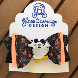May include: A black and orange bow with a white Mickey Mouse ghost charm. The bow has a black and white print with the words "Down Main Street" and "Treat".