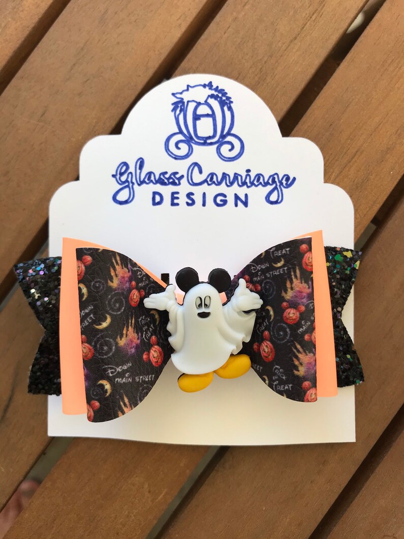 May include: A black and orange bow with a white Mickey Mouse ghost charm. The bow has a black and white print with the words "Down Main Street" and "Treat".