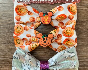 Puede incluir: Tres lazos para el pelo con temática de Halloween. Dos lazos tienen un fondo blanco con calabazas naranjas y una cara de Mickey Mouse con una calabaza en la cabeza. El tercer lazo es plateado con un patrón de fantasmas blancos. Los lazos están dispuestos sobre una superficie de madera con el texto "Glass Carriage Design" encima.
