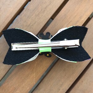 May include: A black and white bow with a green and yellow Mickey Mouse design. The bow is attached to a silver alligator clip.