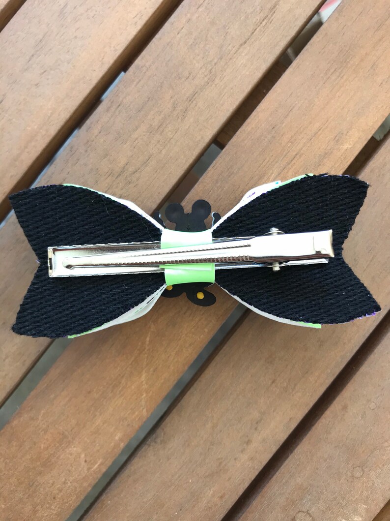 May include: A black and white bow with a green and yellow Mickey Mouse design. The bow is attached to a silver alligator clip.