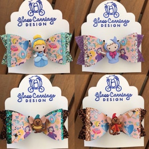 Könnte beinhalten: Vier Haarbänder mit verschiedenen Disney-Prinzessinnen-Charakteren. Jedes Band hat eine rosa Basis mit einem Glitzerüberzug und einen anderen Charakter-Anhänger. Die Charaktere sind Cinderella, Prinz Charming, Gus und Robin Hood. Die Bänder befinden sich alle auf weißen Karten mit dem Text "Glass Carriage Design".