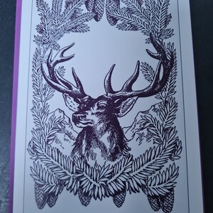 May include: A black and white illustration of a deer's head with antlers framed by a wreath of pine branches and pine cones.