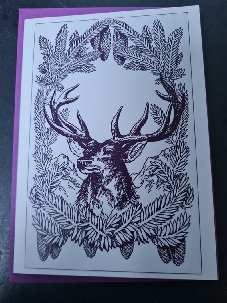 May include: A black and white illustration of a deer's head with antlers framed by a wreath of pine branches and pine cones.