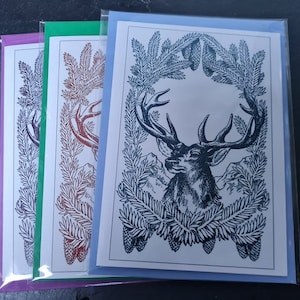 May include: Three greeting cards with a black and white illustration of a deer's head with antlers surrounded by pine branches and pine cones. The cards are in different colors: purple, green, and blue.