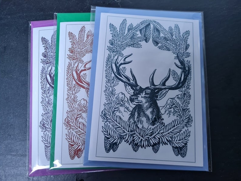 May include: Three greeting cards with a black and white illustration of a deer's head with antlers surrounded by pine branches and pine cones. The cards are in different colors: purple, green, and blue.