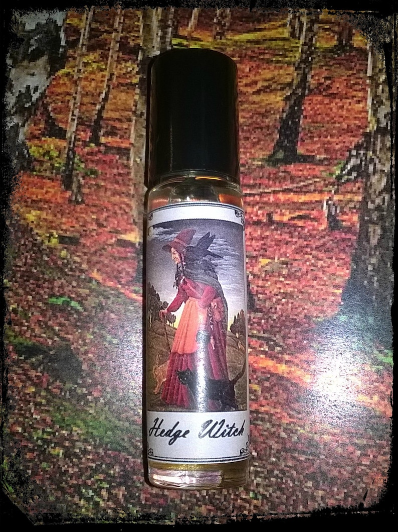 May include: A clear glass roll-on bottle with a black cap and a white label that says "Hedge Witch". The label features an illustration of a woman in a red dress and a black cat walking through a forest.