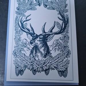 May include: A blue and white illustration of a deer's head with antlers framed by a wreath of pine branches and pine cones.