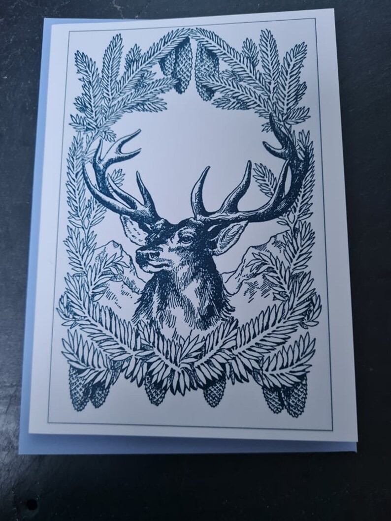 May include: A blue and white illustration of a deer's head with antlers framed by a wreath of pine branches and pine cones.