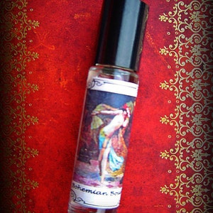 Może przedstawiać: Przezroczysta szklana butelka perfum z czarną zakrętką, z etykietą z ilustracją w stylu vintage kobiety. Butelka umieszczona na teksturowanym czerwonym tle ze złotymi zdobieniami. Etykieta głosi "Bohemian Soul".