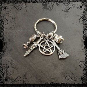 May include: Silver-toned keychain featuring charms of a chalice, sword, pentagram, apple, and broom. The charms are attached to a textured ring. The background is a dark gray surface with decorative borders.