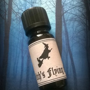 May include: A small glass bottle with a black cap and a white label. The label has a black silhouette of a witch flying on a broomstick with a cat. The text on the label reads "Witch's Flying".