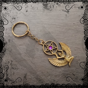 May include: An antique gold-coloured keychain featuring a pentagram with a purple gem and a winged figure. The keychain includes a chain and a circular key ring. The design has a vintage appearance.