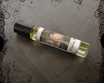 May include: A clear glass roll-on bottle with a black cap and a label that says "Kitchen Witch" and features a painting of a woman in a forest.