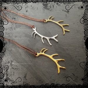 May include: Three antler pendant necklaces. One is silver, and two are gold-toned. Each antler pendant is attached to a brown cord necklace. The pendants are displayed on a dark grey surface.