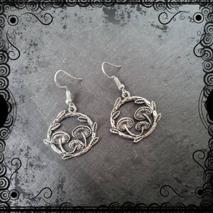 May include: Silver earrings with a circular design featuring three mushroom silhouettes surrounded by leaves.