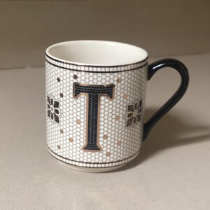May include: White ceramic mug with a black handle and a mosaic tile design. The mug features a large black letter "T" in the center, surrounded by a pattern of small gold and black tiles. The mug is on a light-colored countertop.