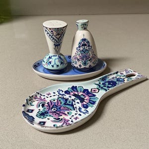 May include: A ceramic salt and pepper shaker set with a matching spoon rest. The set features a blue, purple, and white floral design. The spoon rest is approximately 9 inches long. The salt and pepper shakers are on a small blue plate.