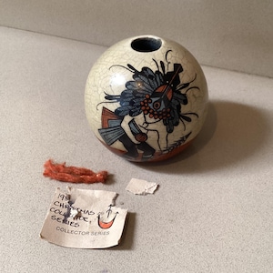 May include: A round, off-white ceramic vase with a detailed Native American design in blue, black, and orange. The vase has a small opening at the top and a cracked glaze. A torn tag and orange string are next to the vase.