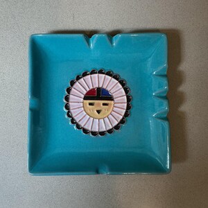 May include: A turquoise square ashtray with a central design featuring a sun face with red, black, and blue accents. The ashtray has four notched areas for resting cigarettes.