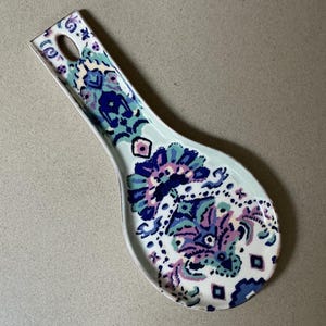 May include: A ceramic spoon rest with a decorative floral pattern in shades of blue, purple, and pink. The spoon rest has a light blue background and a hole in the handle for hanging.