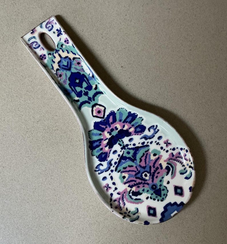 May include: A ceramic spoon rest with a decorative floral pattern in shades of blue, purple, and pink. The spoon rest has a light blue background and a hole in the handle for hanging.