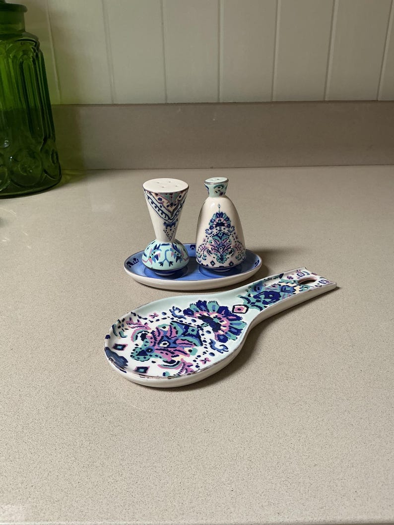 May include: A ceramic salt and pepper shaker set with a matching spoon rest. The set features a blue, purple, and white floral design. The spoon rest is approximately 9 inches long. The salt and pepper shakers are on a small blue plate.