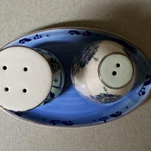 May include: A ceramic salt and pepper set with a blue and white floral design. The set includes two shakers and a matching oval-shaped tray. The shakers have a white top with holes for dispensing the contents.
