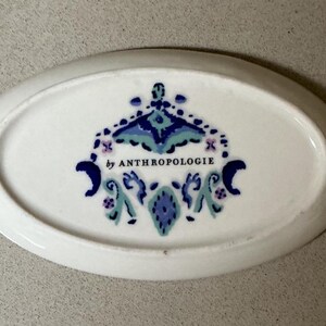 May include: White oval dish with a decorative blue, teal, and purple design. The dish has the text "by ANTHROPOLOGIE" in the center. The dish is a decorative item.