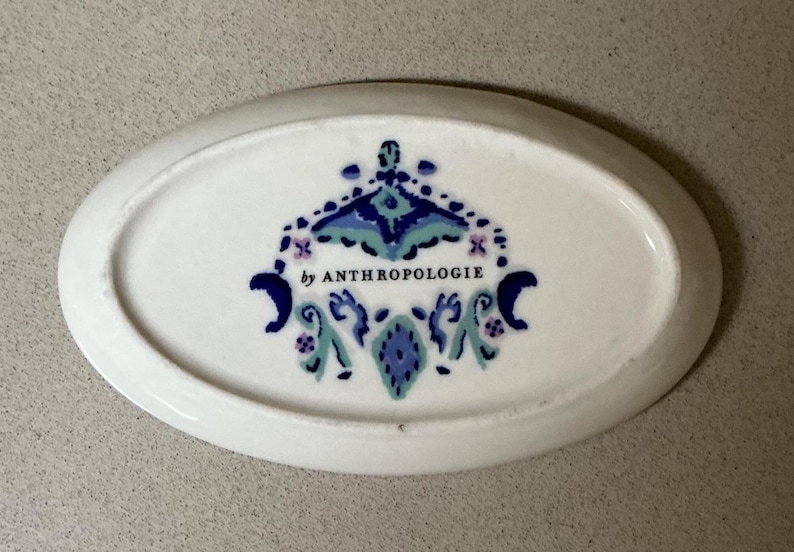 May include: White oval dish with a decorative blue, teal, and purple design. The dish has the text "by ANTHROPOLOGIE" in the center. The dish is a decorative item.