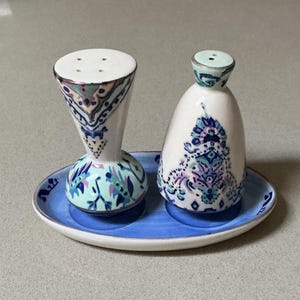 May include: A ceramic salt and pepper set with a blue and white floral design. The set includes a tall, hourglass-shaped salt shaker and a smaller, rounded pepper shaker, both resting on a blue oval tray. The design features intricate patterns in shades of blue, green, and purple.