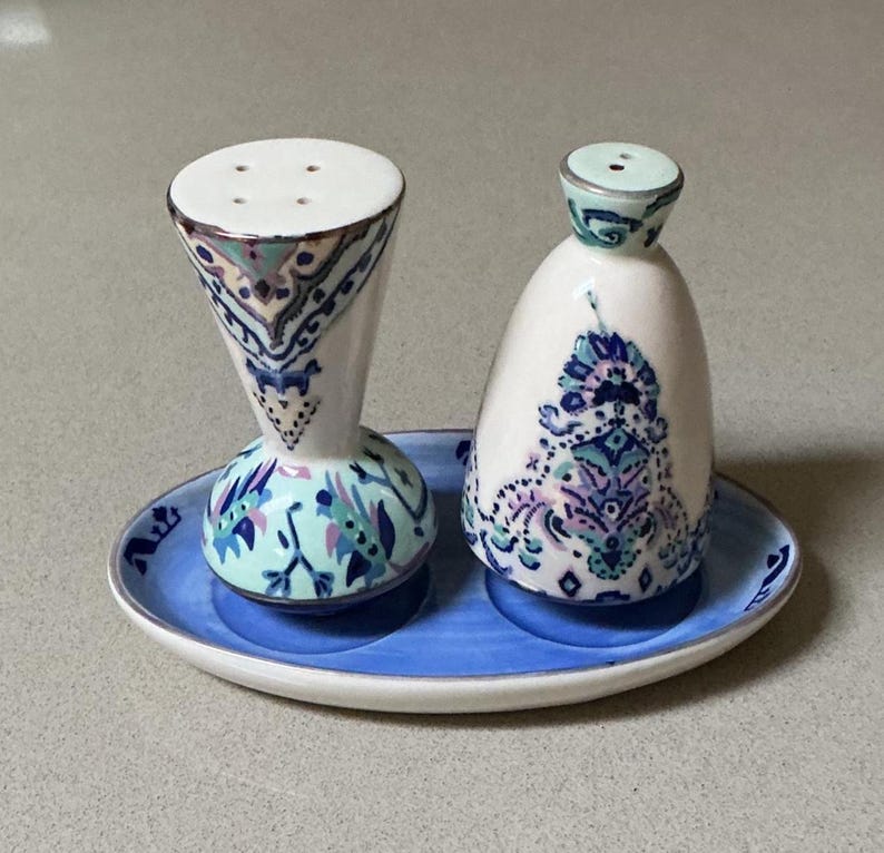 May include: A ceramic salt and pepper set with a blue and white floral design. The set includes a tall, hourglass-shaped salt shaker and a smaller, rounded pepper shaker, both resting on a blue oval tray. The design features intricate patterns in shades of blue, green, and purple.