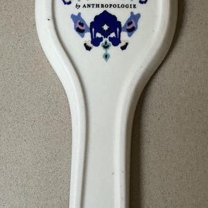 May include: White ceramic spoon rest with a decorative floral design in teal, blue, and pink. The spoon rest is marked "by ANTHROPOLOGIE" and has a handle with a hole for hanging. A kitchen accessory.