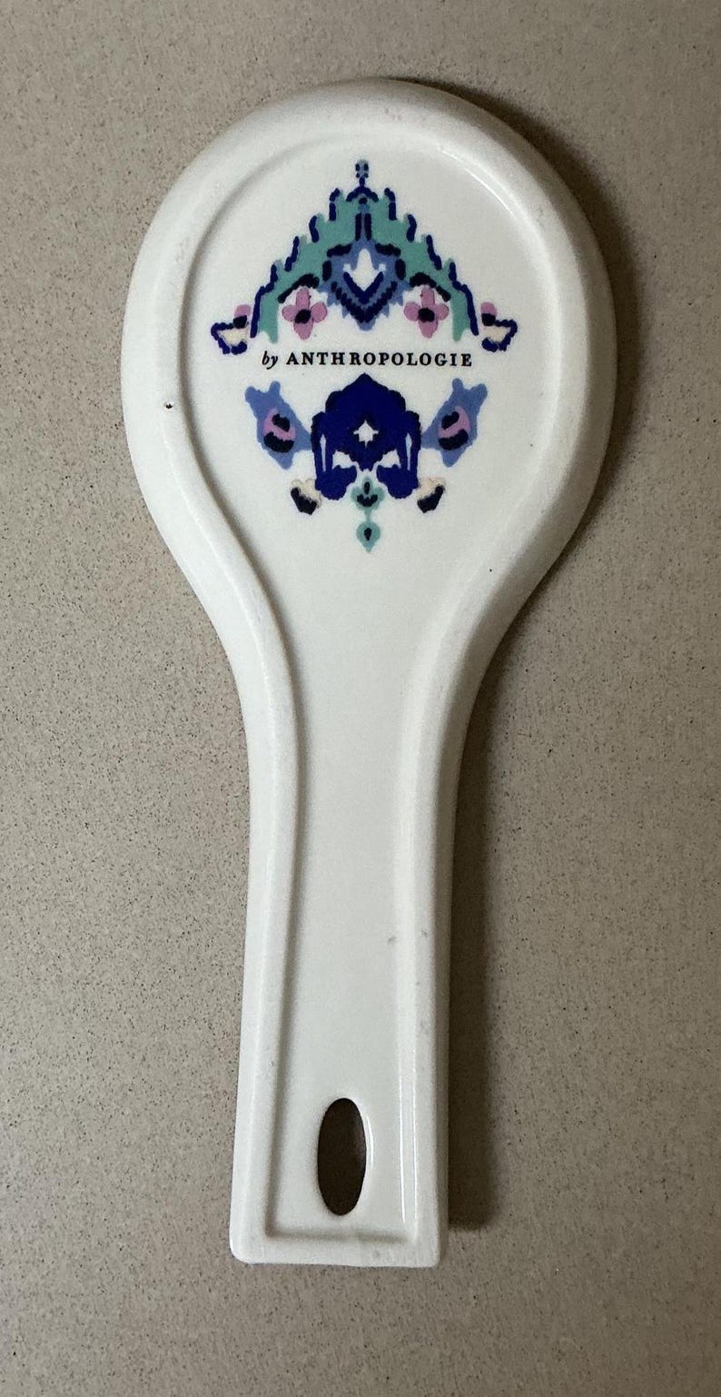 May include: White ceramic spoon rest with a decorative floral design in teal, blue, and pink. The spoon rest is marked "by ANTHROPOLOGIE" and has a handle with a hole for hanging. A kitchen accessory.