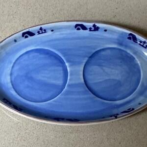 May include: Oval blue ceramic serving dish with two circular indentations. The dish has a white rim and is decorated with dark blue stylized motifs around the edge. The interior is a lighter shade of blue.
