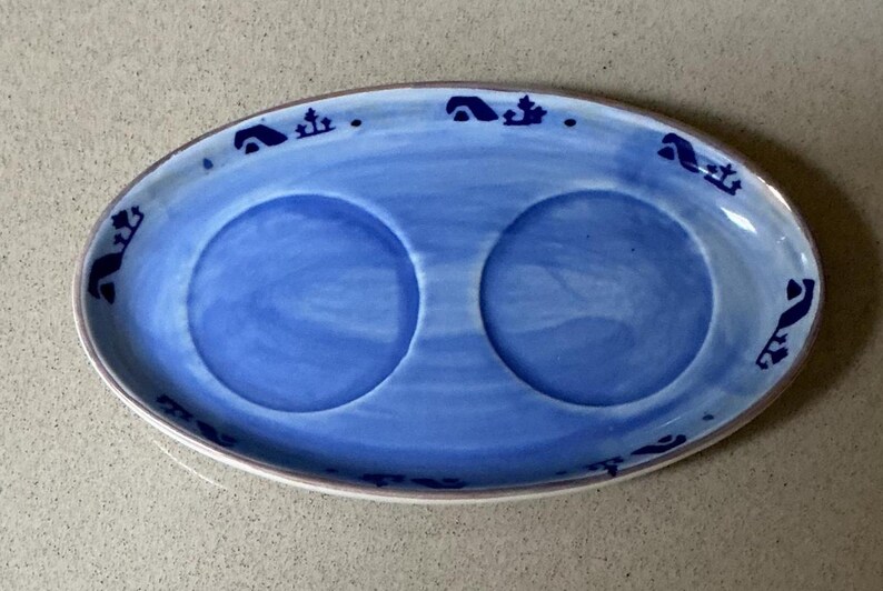 May include: Oval blue ceramic serving dish with two circular indentations. The dish has a white rim and is decorated with dark blue stylized motifs around the edge. The interior is a lighter shade of blue.