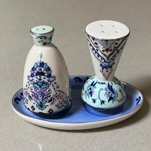 May include: A ceramic salt and pepper set with a matching tray. The shakers are white with blue, purple, and teal floral designs. The tray is light blue with a white rim. The salt shaker is a rounded shape, and the pepper shaker is a cone shape.