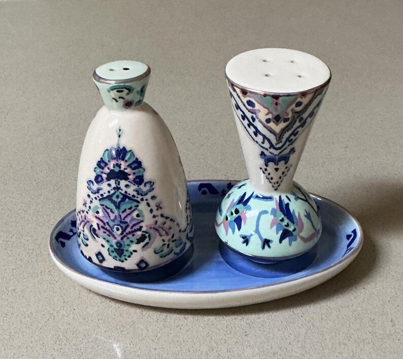 May include: A ceramic salt and pepper set with a matching tray. The shakers are white with blue, purple, and teal floral designs. The tray is light blue with a white rim. The salt shaker is a rounded shape, and the pepper shaker is a cone shape.