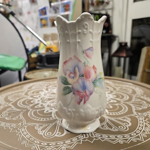 May include: A cream-colored ceramic vase with a scalloped rim and a flared base. The vase is decorated with a floral design in shades of pink, blue, and purple. The vase is approximately 8 inches tall.
