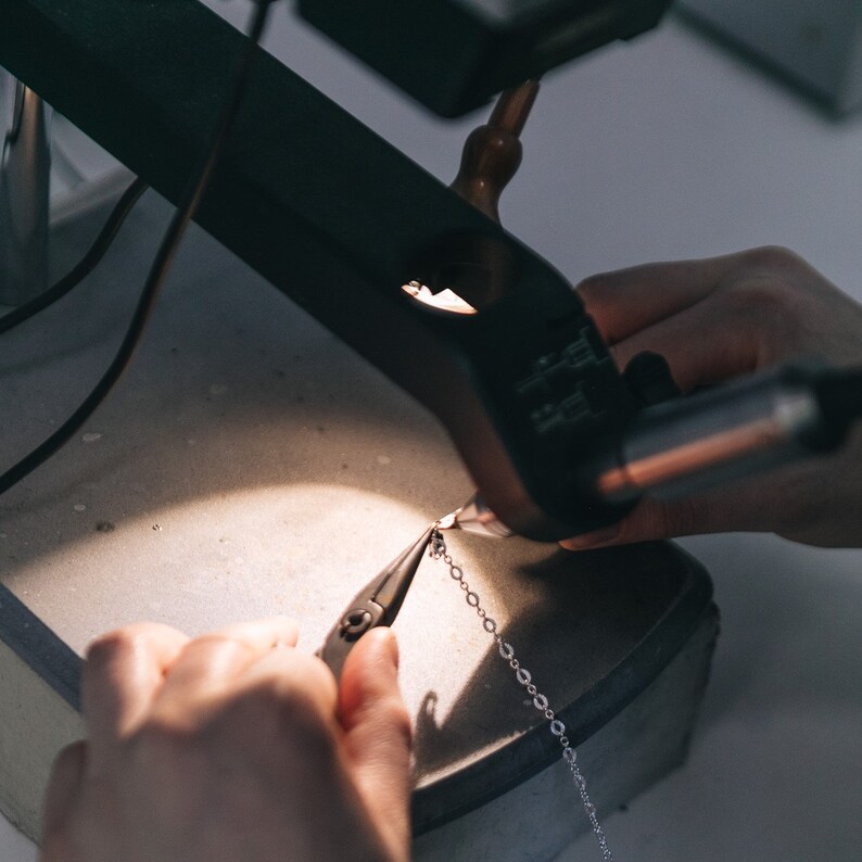 May include: A close-up of a person's hands using pliers to attach a silver chain to a piece of jewelry. The jewelry is being worked on under a magnifying glass.
