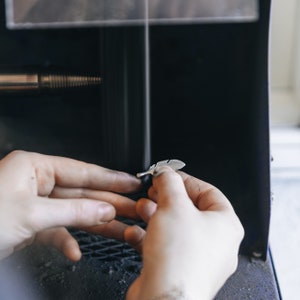 May include: A person's hands holding a silver feather pendant. The pendant is being polished in a machine, creating a cloud of dust.