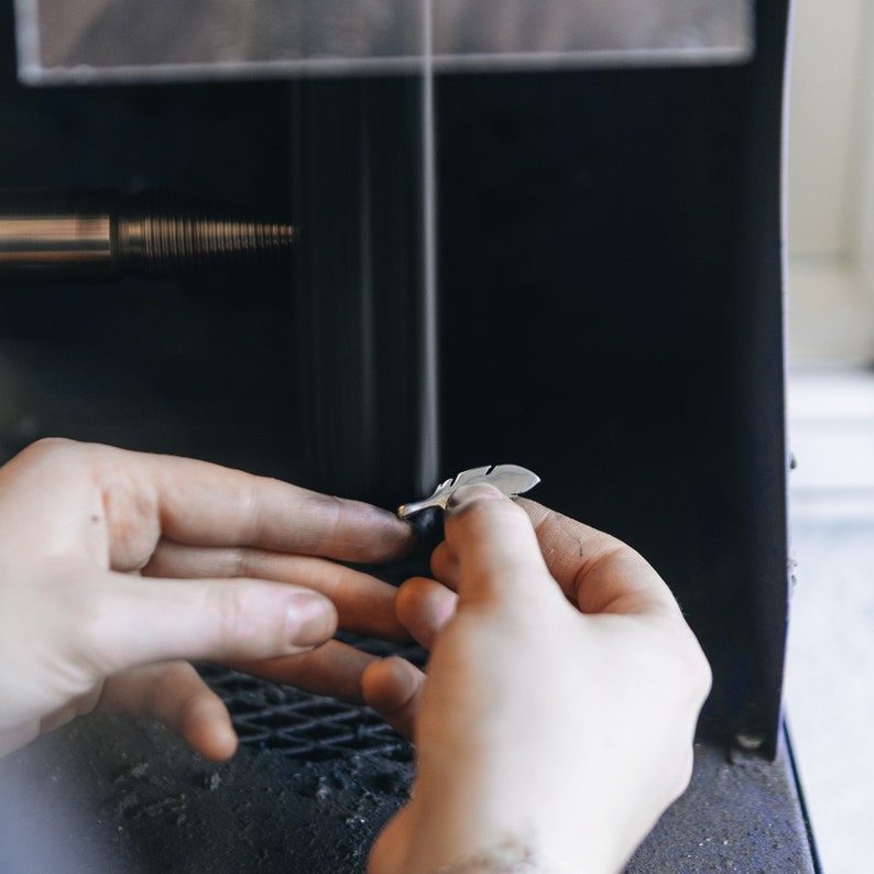 May include: A person's hands holding a silver feather pendant. The pendant is being polished in a machine, creating a cloud of dust.