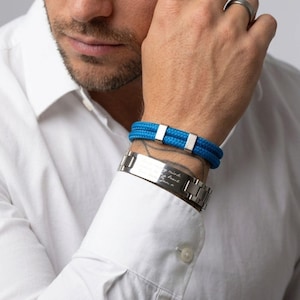 Puede incluir: Primer plano de dos pulseras. Una es una pulsera de cuerda azul con cierres rectangulares plateados. La otra es una pulsera de metal plateado con una placa rectangular con texto grabado. La persona lleva una camisa blanca.