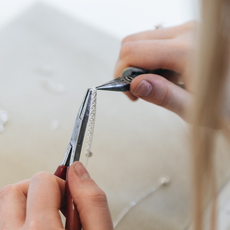 May include: Close-up of a person's hands using pliers to attach a silver chain.